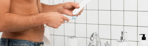 Vista recortada del hombre sin camisa apretando la pasta de dientes en el cepillo de dientes en el baño, pancarta - foto de stock