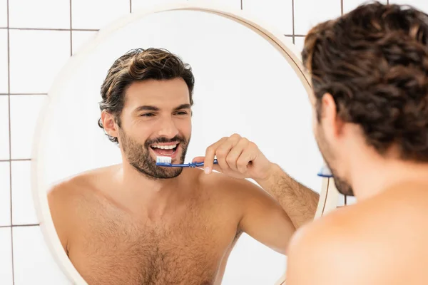 Sonriente hombre sin camisa reflejándose en el espejo mientras se cepilla los dientes en el baño - foto de stock