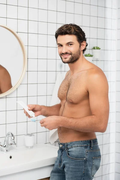 El hombre sin camisa sonriendo a la cámara mientras sostiene la pasta de dientes y el cepillo de dientes en el baño - foto de stock