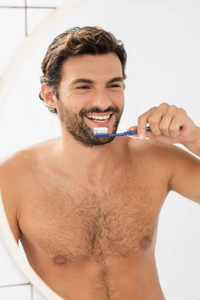 Hombre sin camisa sonriendo al espejo mientras sostiene la pasta de dientes y el cepillo de dientes en el baño - foto de stock