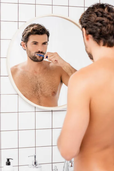 Без сорочки чоловік дивиться на дзеркало під час чищення зубів на розмитому передньому плані у ванній — стокове фото