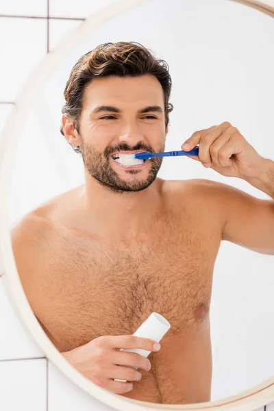 Muscular man holding tube with toothpaste while brushing teeth near mirror — Stock Photo