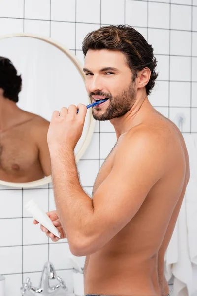 Дерьмовый человек смотрит в камеру, когда чистит зубы и держит трубку с зубной пастой в ванной комнате — стоковое фото