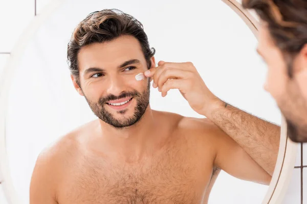 Uomo sorridente che si guarda allo specchio mentre applica la crema per il viso su primo piano sfocato — Foto stock