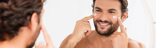 Uomo sorridente che applica crema cosmetica sul viso vicino allo specchio su primo piano sfocato, banner — Foto stock