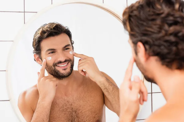 Hombre positivo aplicando crema cosmética en la cara cerca del espejo en primer plano borroso en el baño - foto de stock