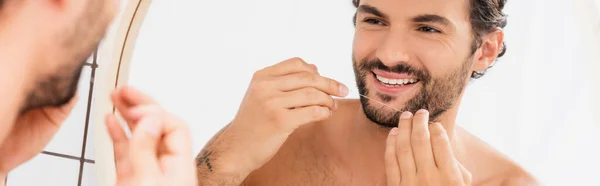 Uomo sorridente che tiene il filo interdentale vicino allo specchio in primo piano sfocato in bagno, banner — Foto stock