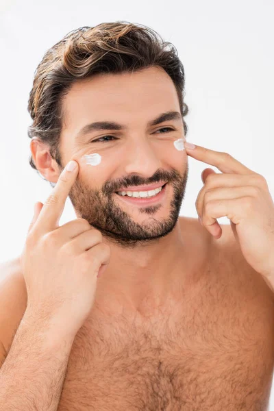 Uomo sorridente senza maglietta che applica crema viso sulle guance isolate sul grigio — Foto stock