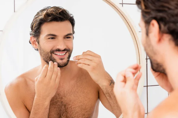 Sonriente hombre sin camisa sosteniendo hilo dental cerca del espejo en el baño - foto de stock