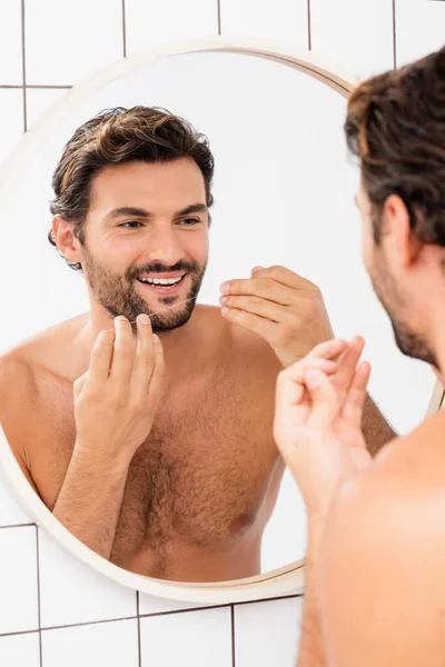 Hombre sin camisa sonriendo mientras sostiene el hilo dental cerca del espejo sobre un fondo borroso - foto de stock
