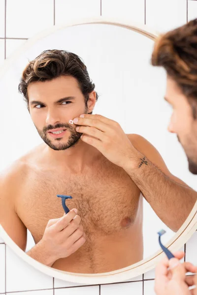 Shirtless man touching cheek with wound and holding razor near mirror on blurred foreground — Stock Photo