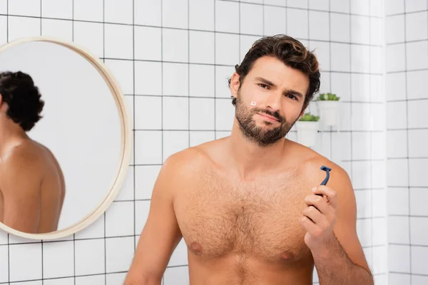 Hombre sin camisa con herida en la mejilla mirando a la cámara mientras sostiene la navaja en el baño - foto de stock