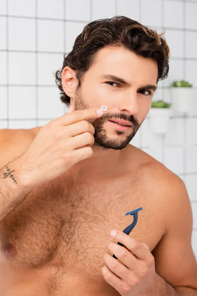 Безсоромний чоловік вказує на рану на щоку, тримаючи бритву у ванній — стокове фото