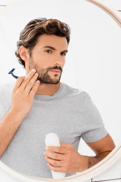 Joven tocando mejilla mientras sostiene espuma de afeitar y afeitadora cerca del espejo - foto de stock