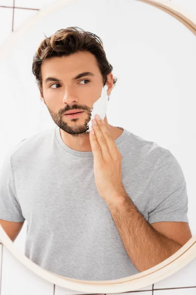 Бородатый мужчина смотрит в зеркало, нанося пену для бритья в ванной комнате — стоковое фото