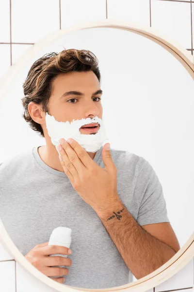 Giovane uomo che applica schiuma da barba vicino specchio in bagno — Foto stock