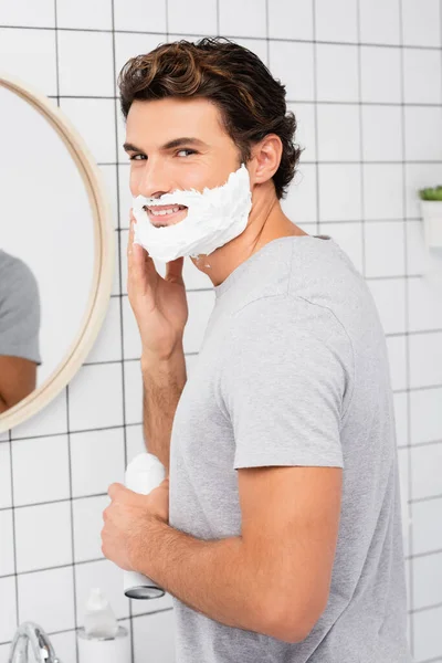 Улыбающийся человек наносит пену для бритья в ванной комнате — стоковое фото