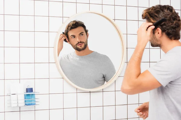 Jeune homme regardant la caméra tout en peignant les cheveux sur le premier plan flou dans la salle de bain — Photo de stock