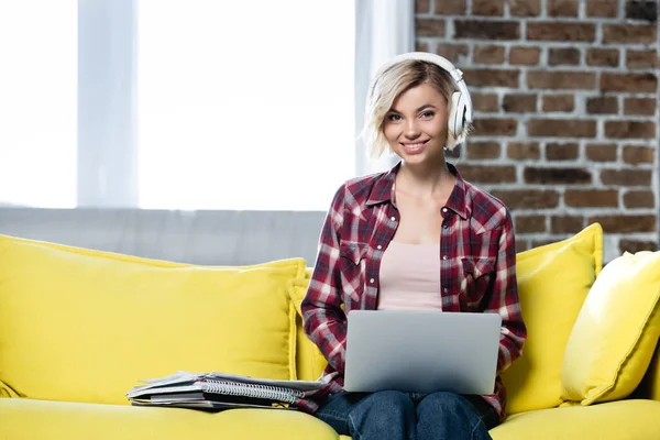 Jeune femme blonde dans un casque assis sur un canapé jaune avec ordinateur portable sur les genoux — Photo de stock