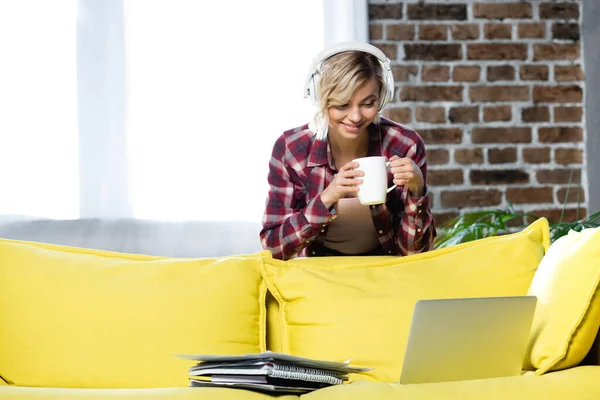 Junge blonde Frau mit Kopfhörer lehnt auf Sofa und trinkt Tee — Stockfoto