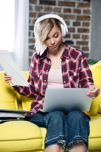 Giovane donna bionda in cuffia guardando il computer portatile e in possesso di documenti — Foto stock