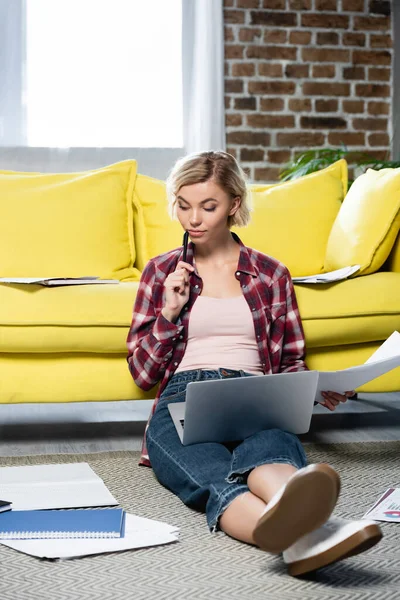 Jeune femme blonde assise sur le sol avec ordinateur portable sur les genoux et travaillant à la maison — Photo de stock