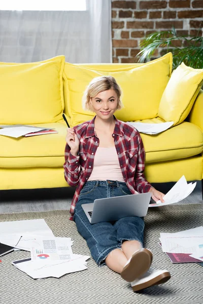 Joven mujer rubia sentada en el suelo con el ordenador portátil en las rodillas y la celebración de documentos - foto de stock
