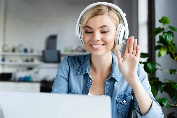 Jeune femme blonde dans les écouteurs ayant conférence en ligne et agitant la main — Photo de stock
