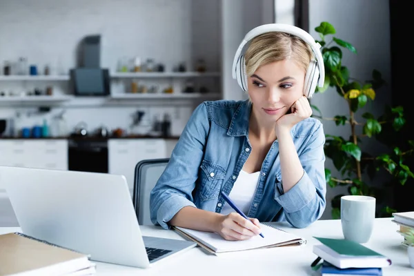 Junge blonde Frau mit Kopfhörer lernt online und macht sich Notizen — Stockfoto