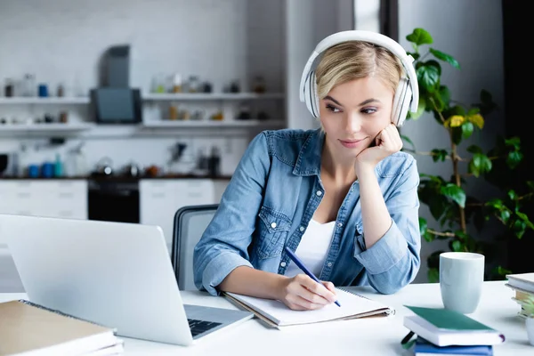 Joven rubia en auriculares estudiando en línea y tomando notas - foto de stock