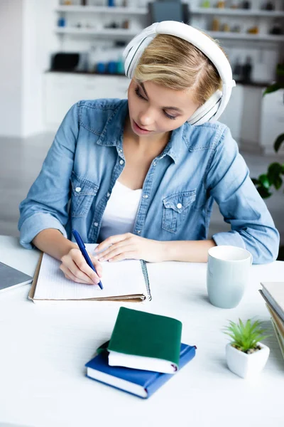 Jeune femme blonde dans les écouteurs étudier en ligne et prendre des notes — Photo de stock