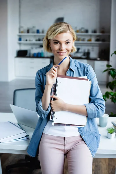 Joven rubia sosteniendo cuadernos y bolígrafo en casa - foto de stock