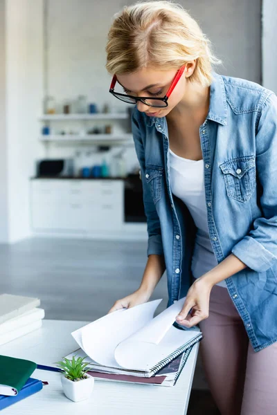 Mujer rubia joven en gafas que trabajan desde casa y pasar páginas de cuaderno - foto de stock