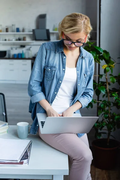 Молода блондинка в окулярах працює з дому, сидячи на столі — Stock Photo