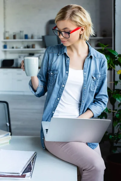 Young blonde woman in eyeglasses working from home sitting on table and drinking tea — Stock Photo