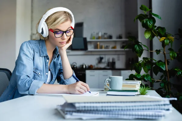 Junge blonde Frau mit Brille und Kopfhörer, die Notizen in Notizbuch macht — Stockfoto