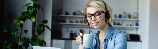 Young blonde woman in eyeglasses and headphones drinking tea, horizontal banner — Stock Photo