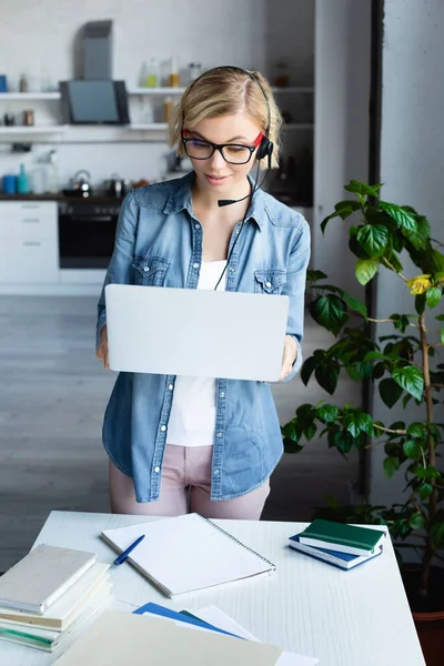 Mujer rubia joven en gafas que trabajan desde casa y la celebración de la computadora portátil - foto de stock
