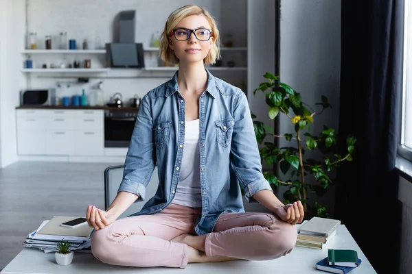 Young blonde woman in eyeglasses sitting in lotus pose on table — Stock Photo