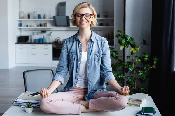 Smiling woman in eyeglasses sitting in lotus pose on table — Stock Photo