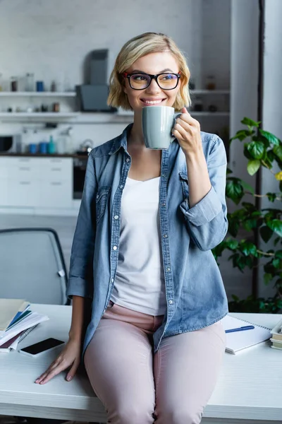 Smiling blonde woman in eyeglasses drinking tea at home — Stock Photo