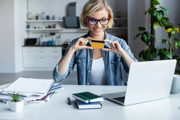 Young blonde woman in eyeglasses holding credit card and making purchase online — Stock Photo