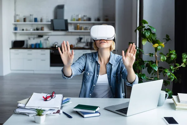 Donna bionda in auricolare vr gesticolare mentre si siede a tavola — Foto stock