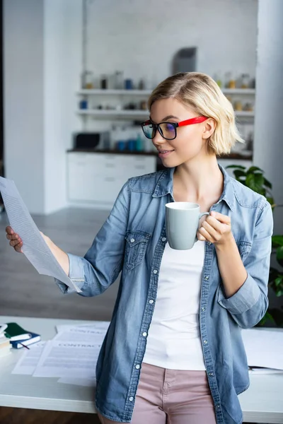 Junge blonde Frau mit Brille schaut sich Dokument an und trinkt Tee — Stockfoto