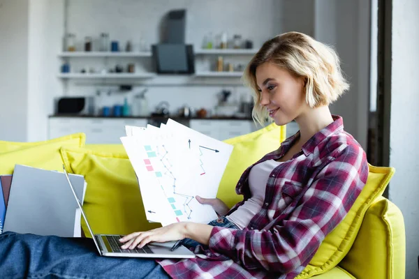 Blonde woman in checkered shirt typing on laptop and holding documents — Stock Photo