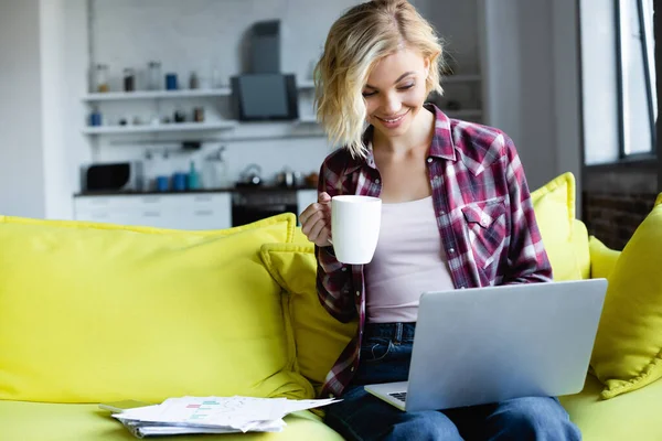 Blonde woman in checkered shirt working from home and drinking tea — Stock Photo