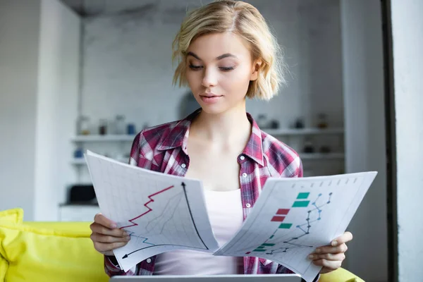 Femme blonde en chemise à carreaux travaillant à la maison et regardant des graphiques — Photo de stock