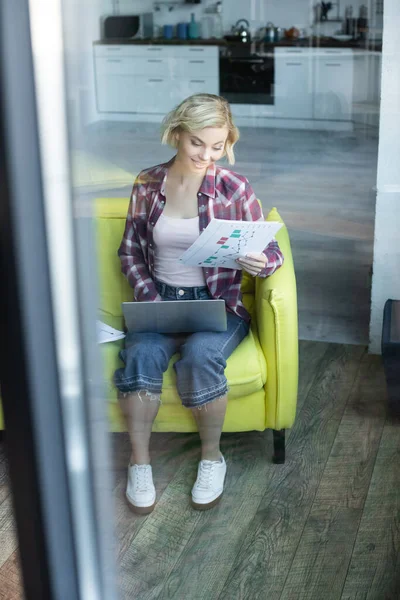 Blick durch das Glas einer blonden Frau im karierten Hemd, die von zu Hause aus arbeitet und Dokumente durchsieht — Stockfoto