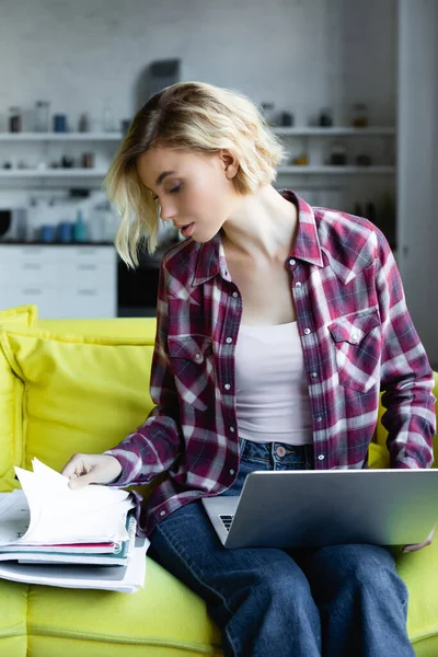 Молода блондинка в картатій сорочці тримає ноутбук і дивиться в документах — стокове фото