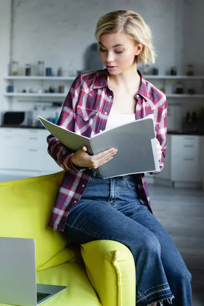 Junge blonde Frau im karierten Hemd auf Sofa sitzend und Dokumente einsehend — Stockfoto